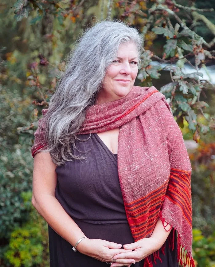 A woman with long gray hair wearing a dark brown dress and a rust-red patterned shawl, standing outdoors in front of a blurred green and brown foliage background.