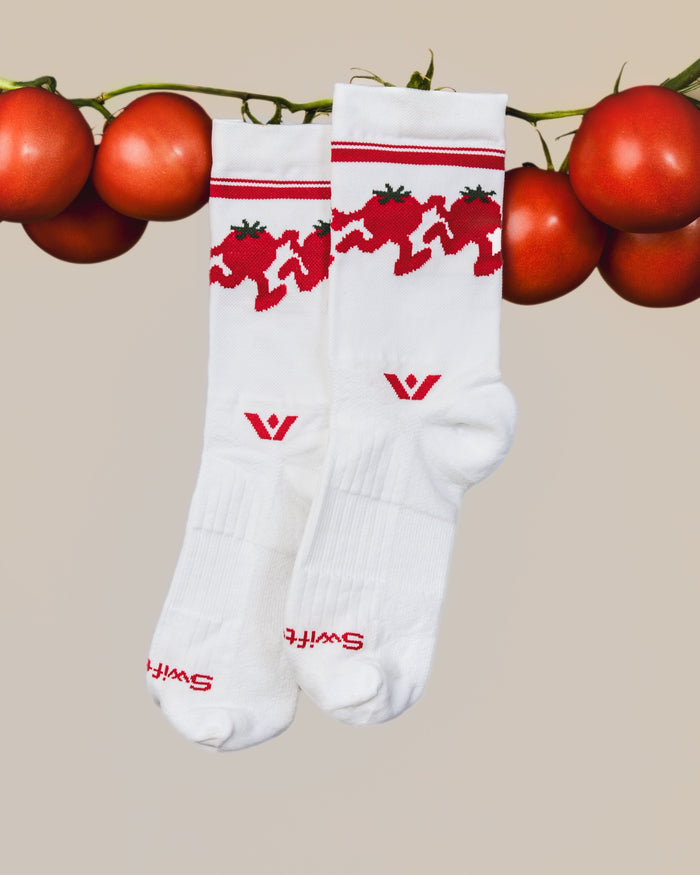 Two white Swiftwick Vision Mid-Crew socks with a red dancing-tomato cuff pattern hanging from a vine of fresh tomatoes.