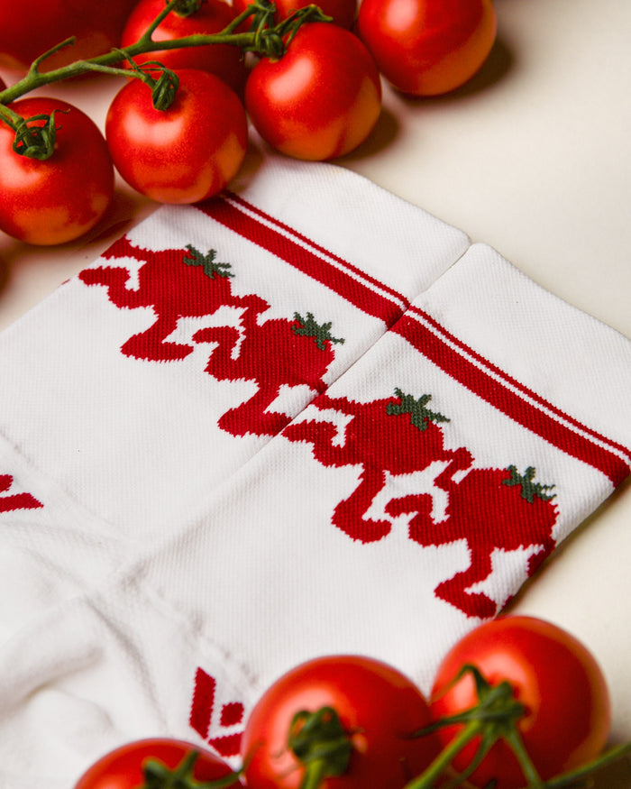 Close-up of white Swiftwick Vision Mid-Crew socks with a red dancing-tomato cuff pattern laid beside fresh red tomatoes.