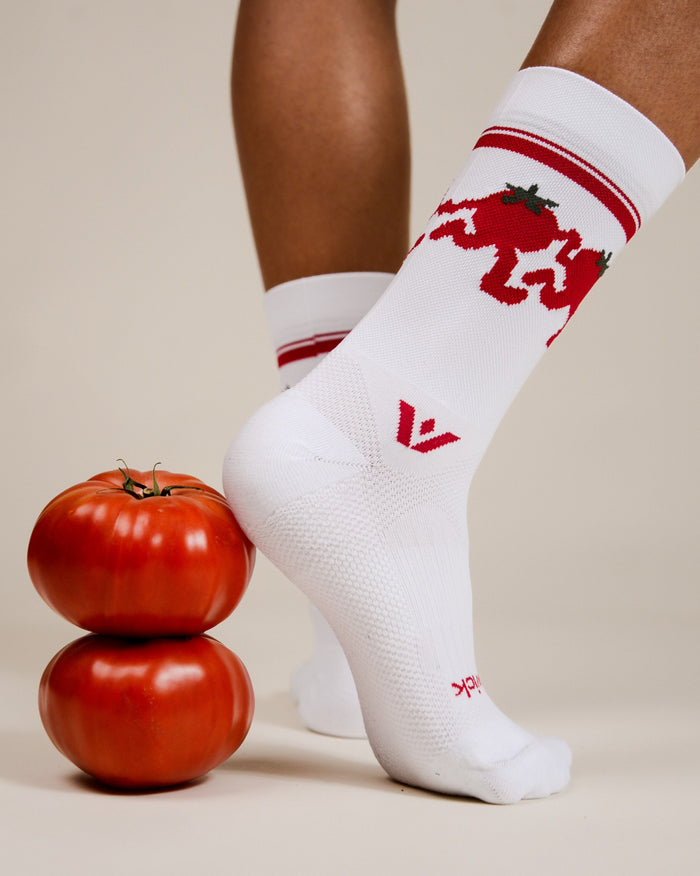 Person wearing white Swiftwick Vision Mid-Crew socks with a red dancing-tomato pattern, standing beside two stacked ripe tomatoes.