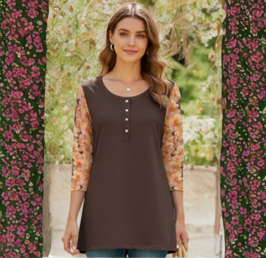 A woman with long wavy brown hair stands centered against a soft-focus garden backdrop. She is wearing a chocolate brown, long-line tunic featuring a round neckline with a short button-down placket and three-quarter length floral print sleeves in shades of peach and orange.