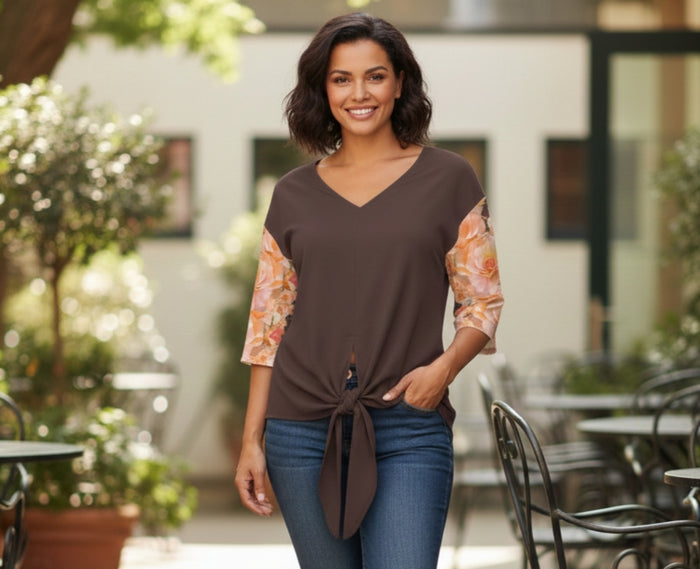 Woman wearing the Apricot Bloom Essential blouse by TimeReproofPortraits, featuring a dark earth-tone bodice, apricot floral print sleeves, and a centered tie-front knot at the waist, styled with blue jeans in an outdoor cafe setting.