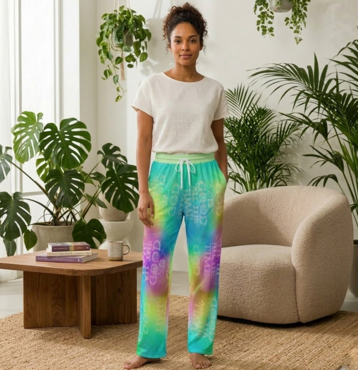 Woman wearing "God is Good" tie-dye lounge pants with open bottoms, made from recycled polyester. Colorful teal and purple print in a bright living room.