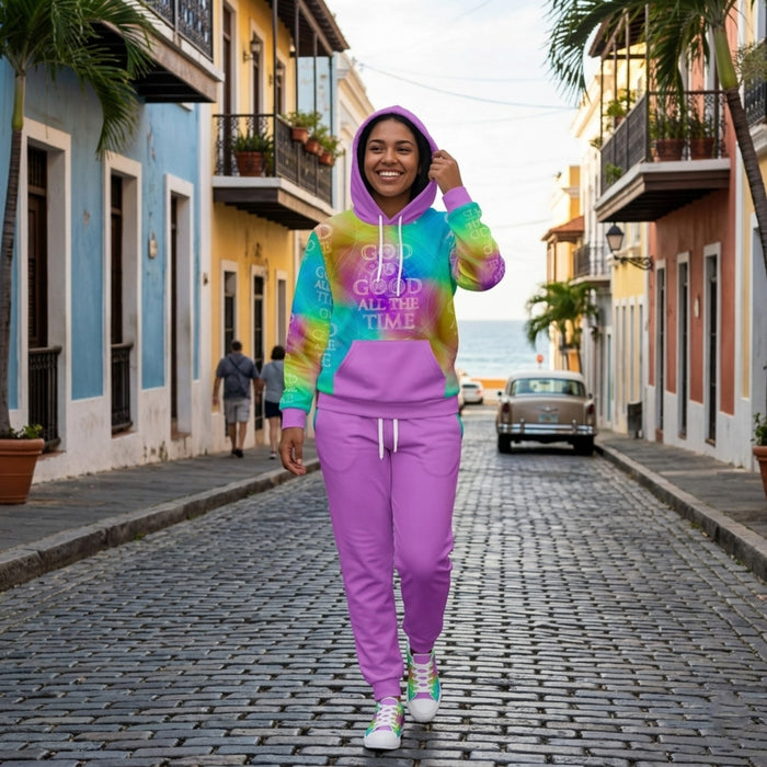A smiling woman walks down a colorful cobblestone street wearing a purple sweatsuit with a vibrant, tie-dye "God is Good All the Time" hoodie.
