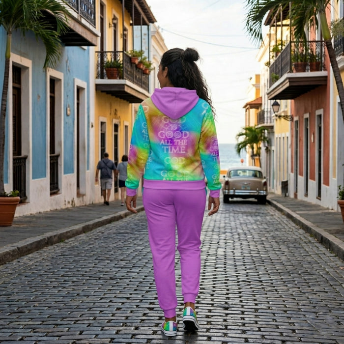 Back view of a woman walking on a cobblestone street wearing a purple sweatsuit and a tie-dye hoodie featuring the text "God is Good All the Time.