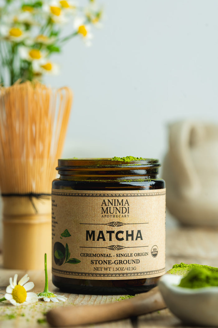 Jar of Anima Mundi Matcha on a wooden surface with flowers and a whisk in the background