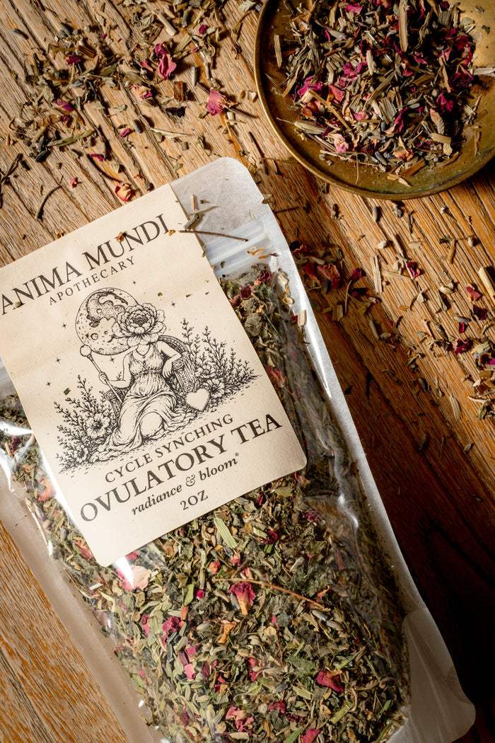A clear plastic bag of Anima Mundi Apothecary Cycle Syncing Ovulatory Tea filled with dried herbs and pink petals, placed on a rustic wooden surface with a small dish of loose tea nearby.