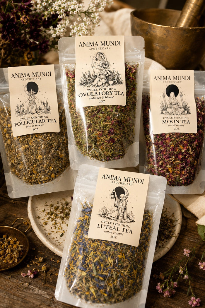 Three clear stand-up pouches of Anima Mundi botanical tea arranged on a rustic wooden surface with floral props and brass kitchenware. The visible labels read Extra Virgin Ovulatory Tea, Follicular Tea, and Luteal Tea.