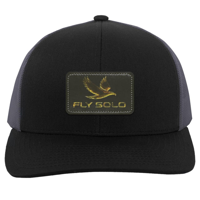 Front product shot of the black trucker hat with grey mesh panels and the signature gold brand logo.