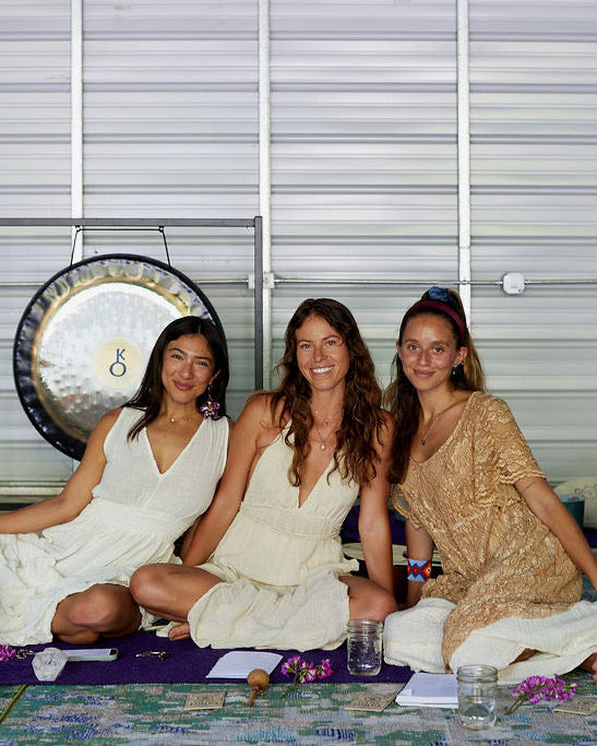 Three women sitting together in a studio setting with a large gong on the left, wearing loose, flowing dresses in white and beige tones. A patterned rug, small mallets, and glass jars are on the floor in front of them, with a white slatted wall behind them.